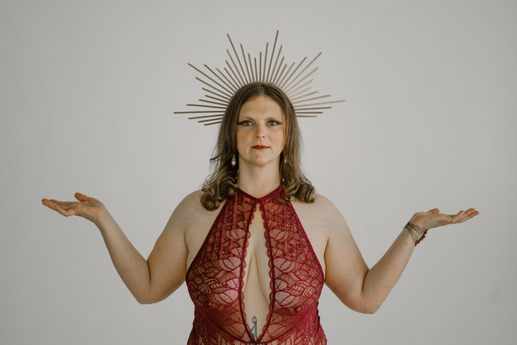 A woman poses in front of a backdrop with her hands open to the sky in the pose of a goddess. She is wearing a gold sun beam crown and lacy lingerie.